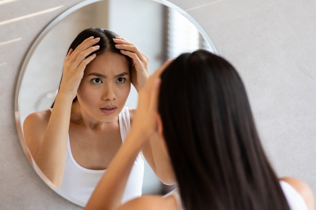 A woman with long hair looks concerned while examining her scalp in a circular bathroom mirror, contemplating the benefits of PRP injections.