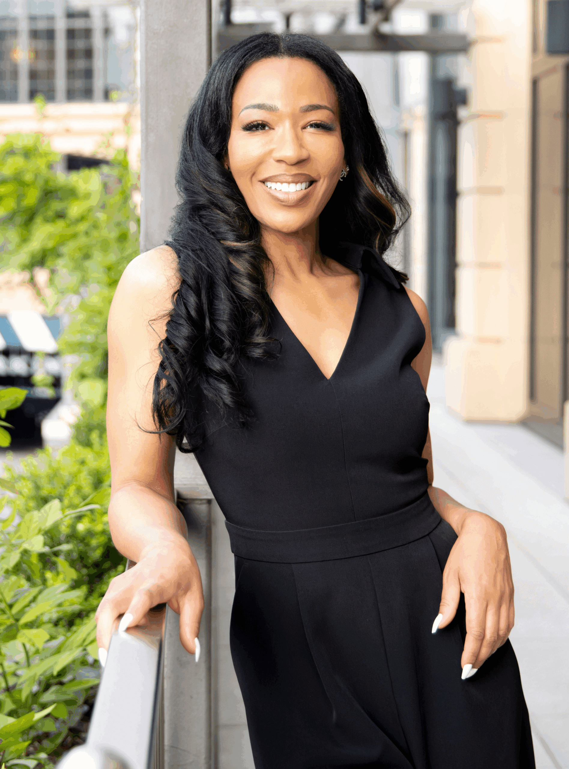 Woman with long black hair wearing a sleeveless black dress, standing outdoors and smiling while leaning on a railing, reminiscent of a scene from Dr. Scott's latest collection.
