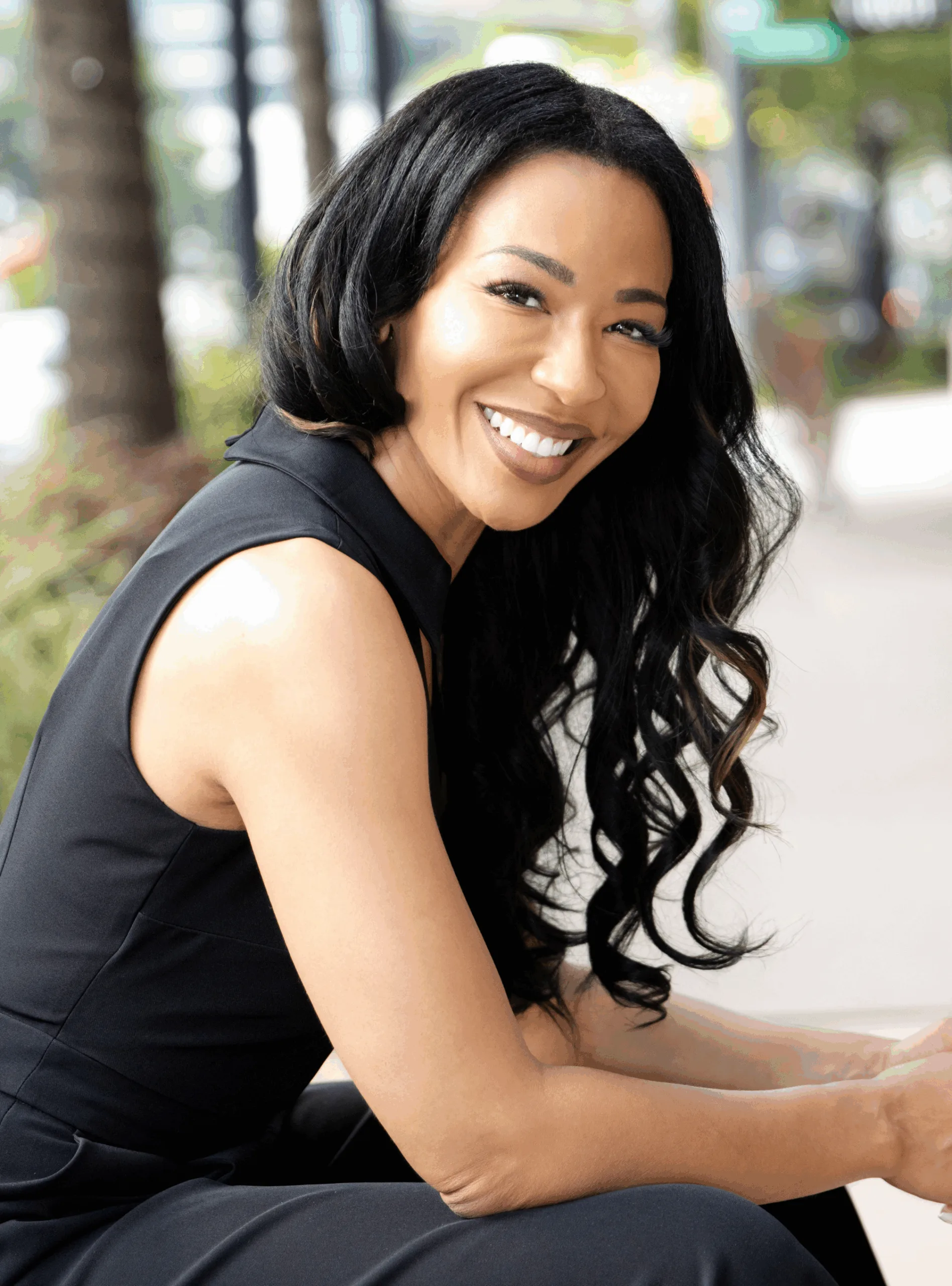 Dr. Scott, a woman with long, wavy black hair wearing a sleeveless black outfit, sits outdoors, smiling at the camera.
